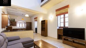 A modern living room with a sofa, a coffee table, and a TV on a wooden stand. There is a kitchen area with cabinets and a window.