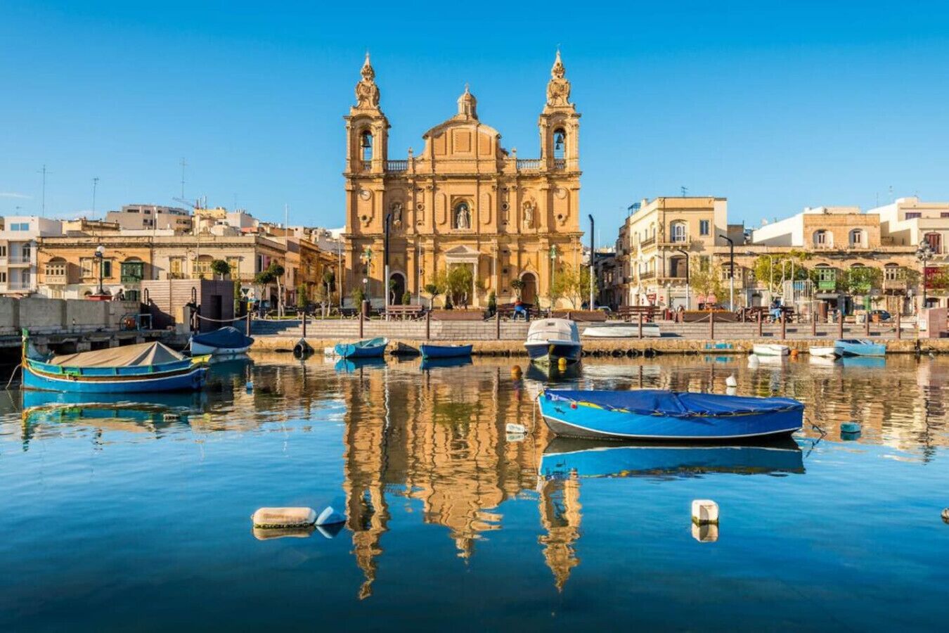 Una banchina con un imponente edificio storico caratterizzato da due torri e una cupola centrale, circondato da acque calme e diverse piccole imbarcazioni.