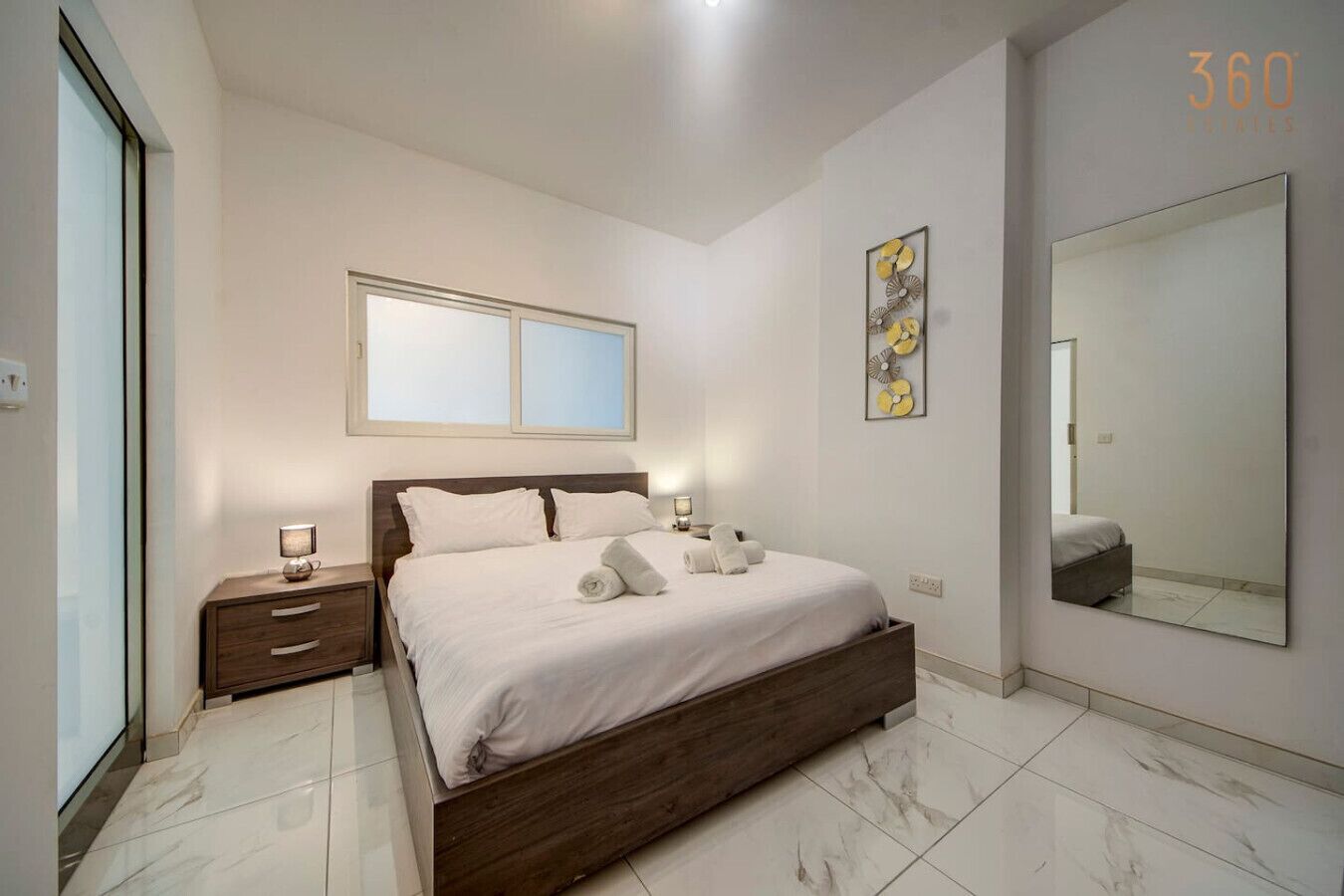 Una camera da letto moderna con un letto in legno, biancheria da letto bianca, un comodino, un tavolino e uno specchio grande, oltre a un’opera d’arte a parete.