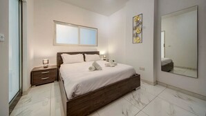 Ein modernes Schlafzimmer mit einem hölzernen Bett, weißer Bettwäsche, einem Nachttisch, einem Beistelltisch, einem großen Spiegel und einem Wandbild.