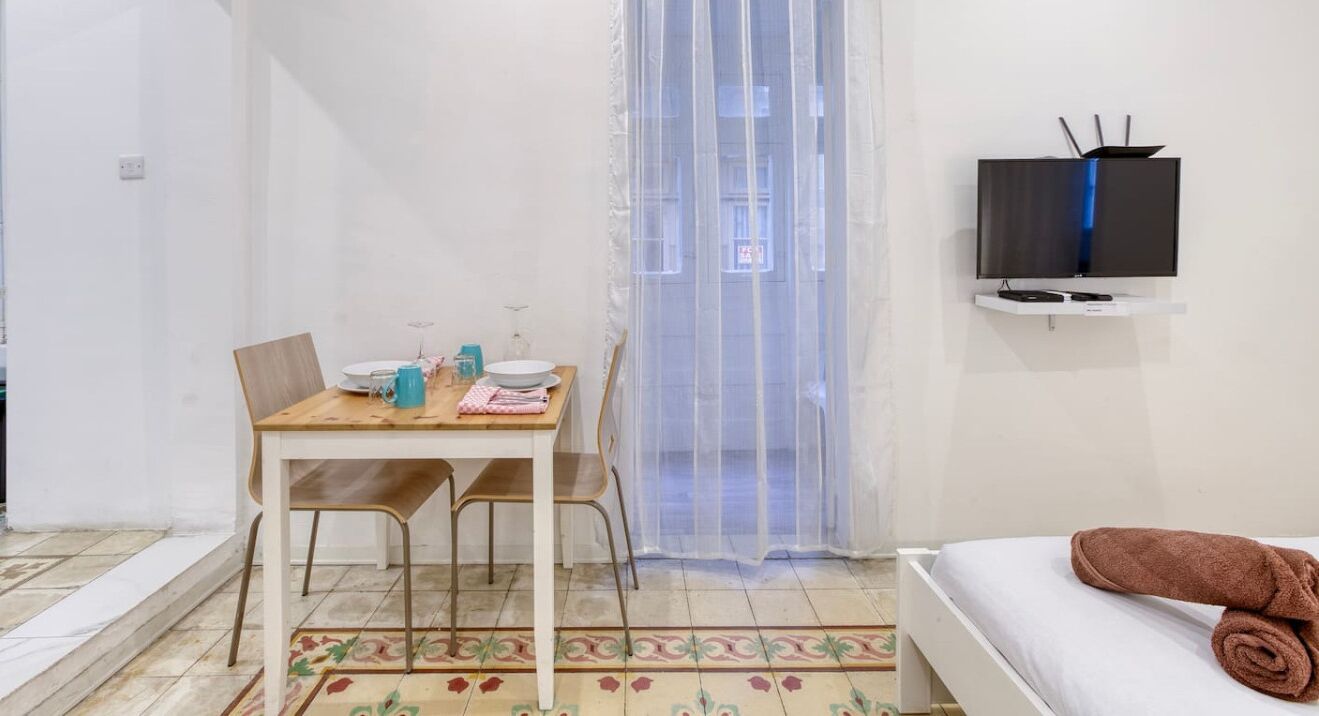 A small, clean room with a bed, a table with chairs, a TV mounted on the wall, and a window with a sheer curtain.
