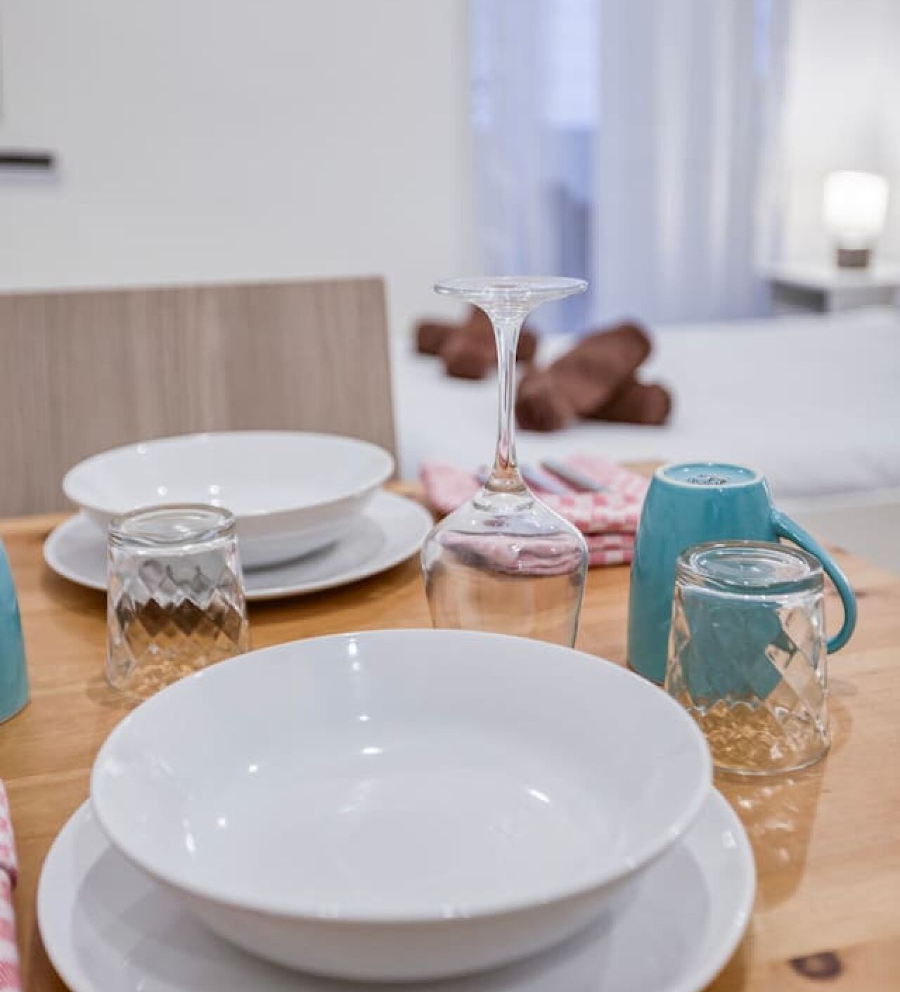 Een eettafel gedekt met witte borden, een helder glas, een blauwe mok en een glazen pot.