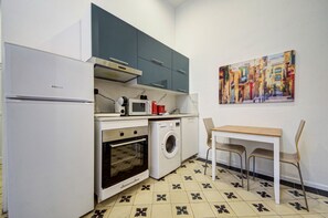 Cucina compatta dotata di frigorifero, forno, microonde e lavatrice. È presente un tavolo con due sedie e un quadro appeso al muro.