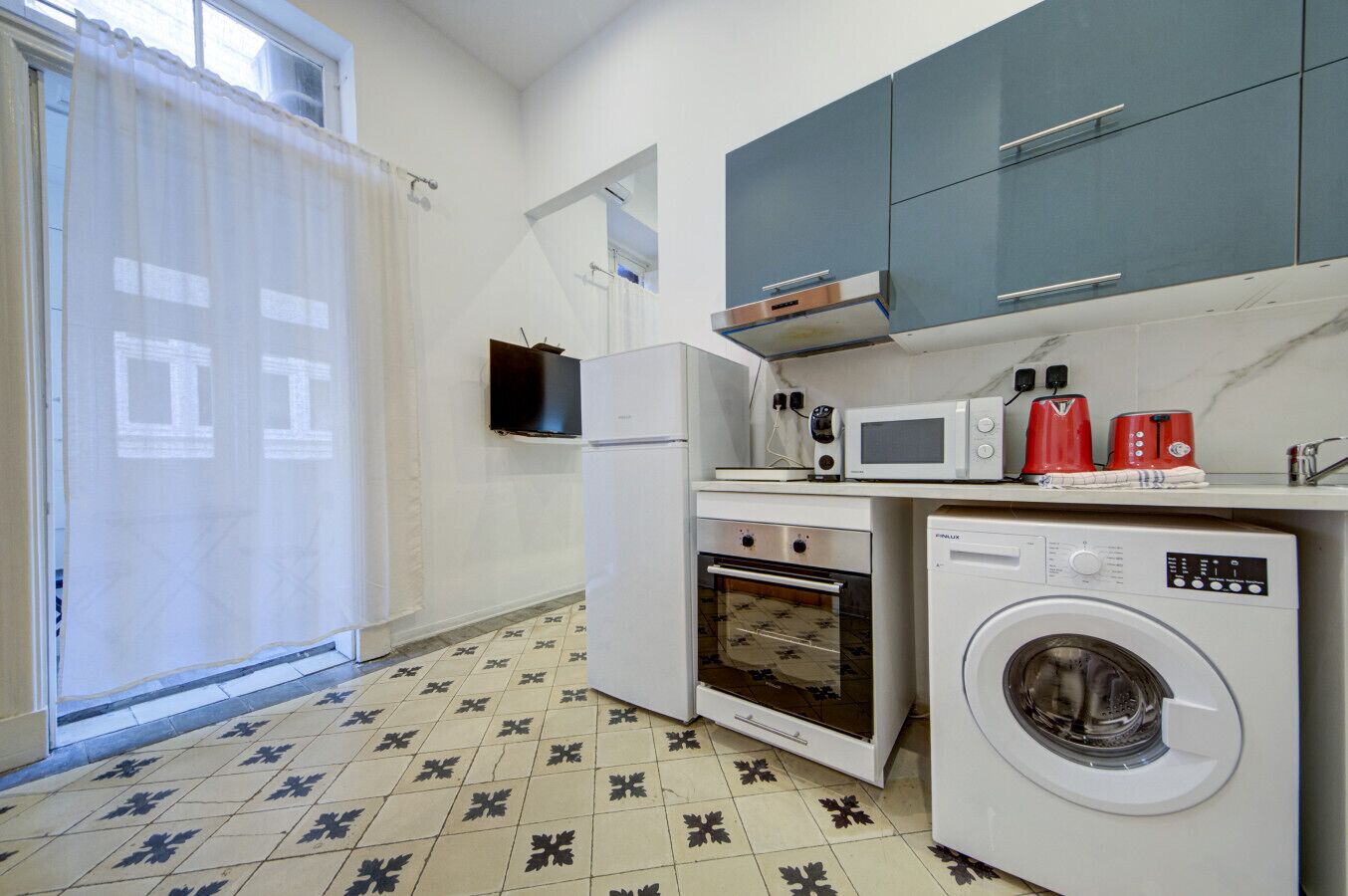 A compact kitchen with a washing machine, microwave, oven, and a small countertop with a toaster and kettle.