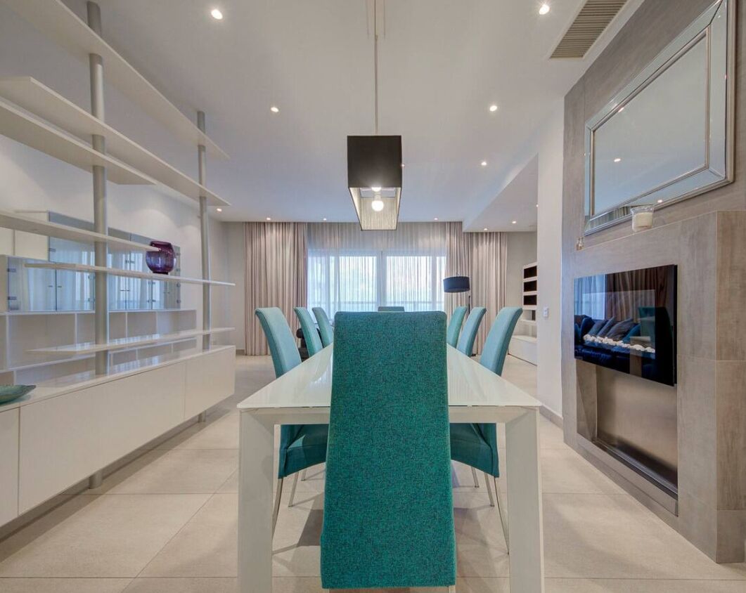 Un espace de salle à manger moderne avec une longue table, des chaises de couleur turquoise et un chemin de table central, également turquoise. À gauche, on trouve des étagères remplies de divers objets, et à droite, un téléviseur à écran plat est fixé au mur.