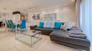 A modern living room with a glass coffee table, a sofa with multiple cushions, and a dining area with chairs and a table.