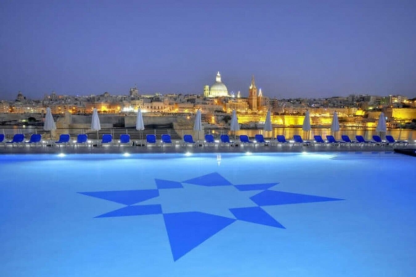 A swimming pool with a star design, blue lounge chairs, and a cityscape with illuminated buildings in the background.