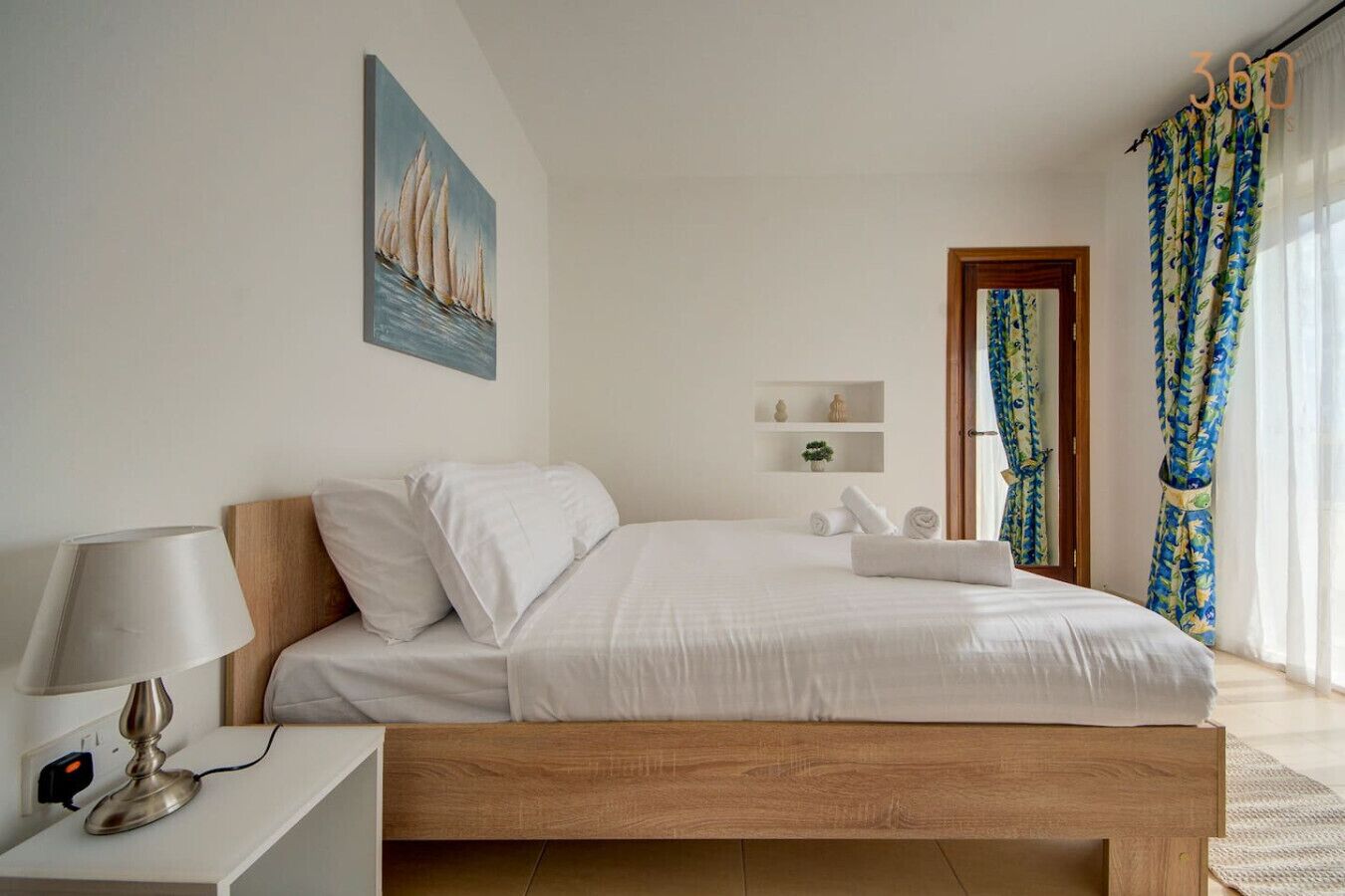 A bedroom with a wooden bed, white bedding, a bedside table with a lamp, a painting on the wall, and a window with curtains.