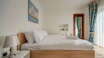Une chambre à coucher avec un lit en bois, du linge de lit blanc, une table de chevet avec une lampe, un tableau au mur et une fenêtre avec des rideaux.