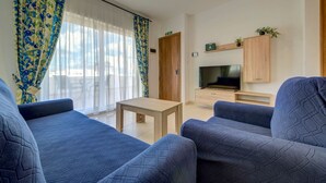 A modern living room with blue patterned sofas, a wooden coffee table, a TV on a wooden stand, and a door with an emergency exit sign.