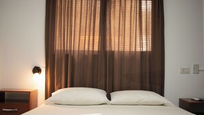 A neatly made bed with white linens and two pillows, a bedside table with a lamp, and a window with brown curtains. - Hotel del Monumento (Salta)