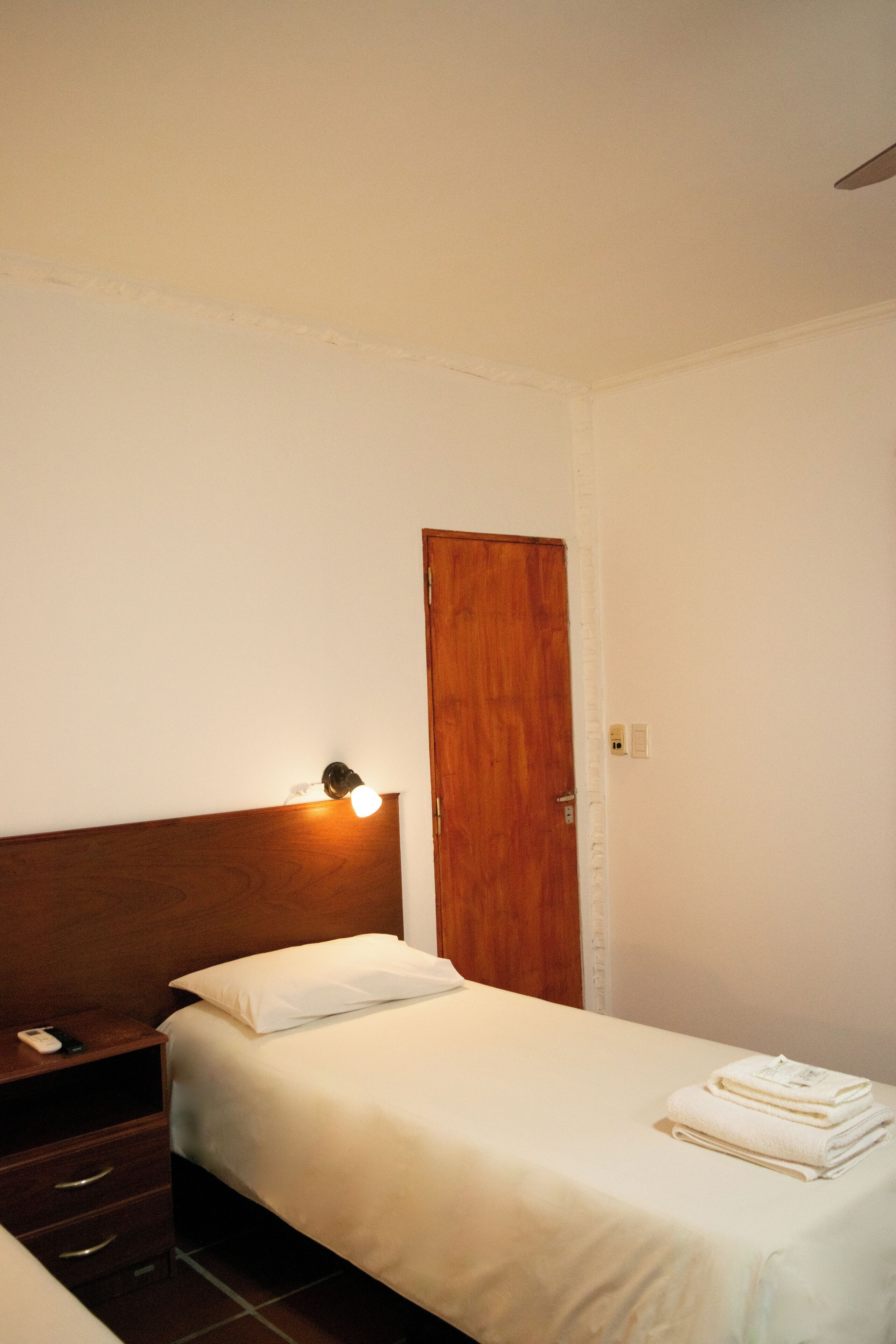 A hotel room with a wooden headboard, a bedside table with a lamp, a bed with white linens, and a door.