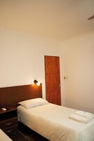 Una habitación de hotel con cabecera de madera, una mesita de noche con lámpara, cama con ropa blanca y una puerta.