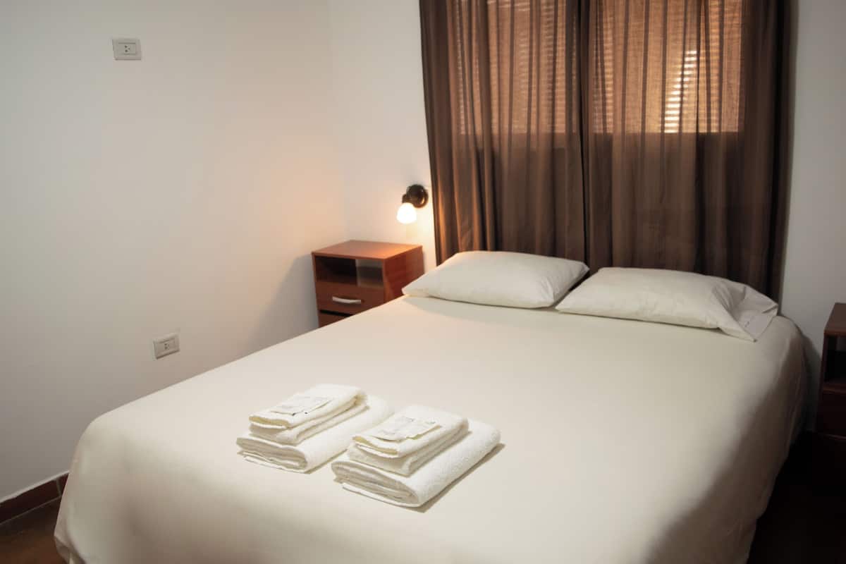 A neatly made bed with two folded towels, a bedside table with a lamp, and a window with curtains.
