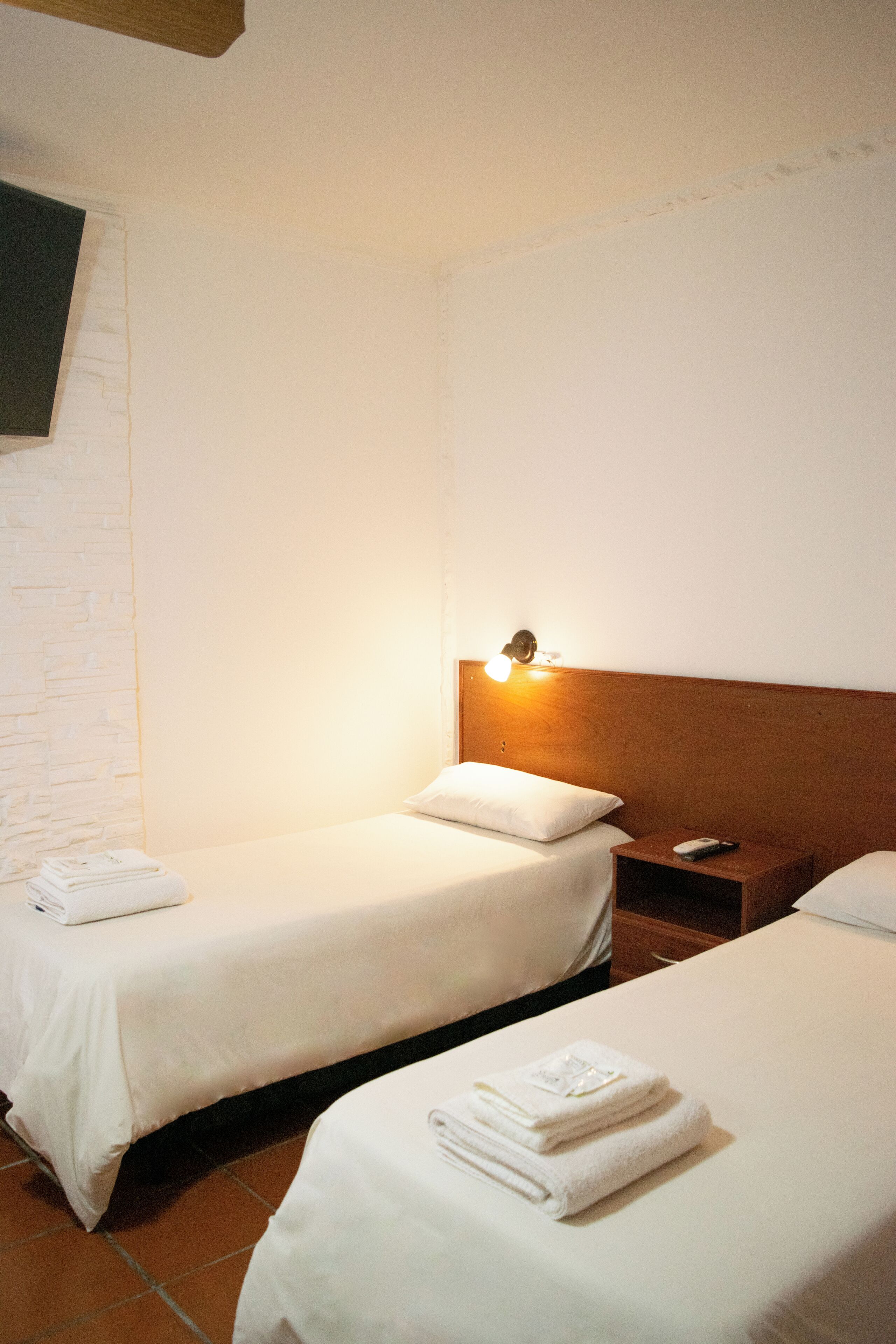 Habitación de hotel con dos camas, cabecero de madera, mesita de noche y televisor montado en la pared.