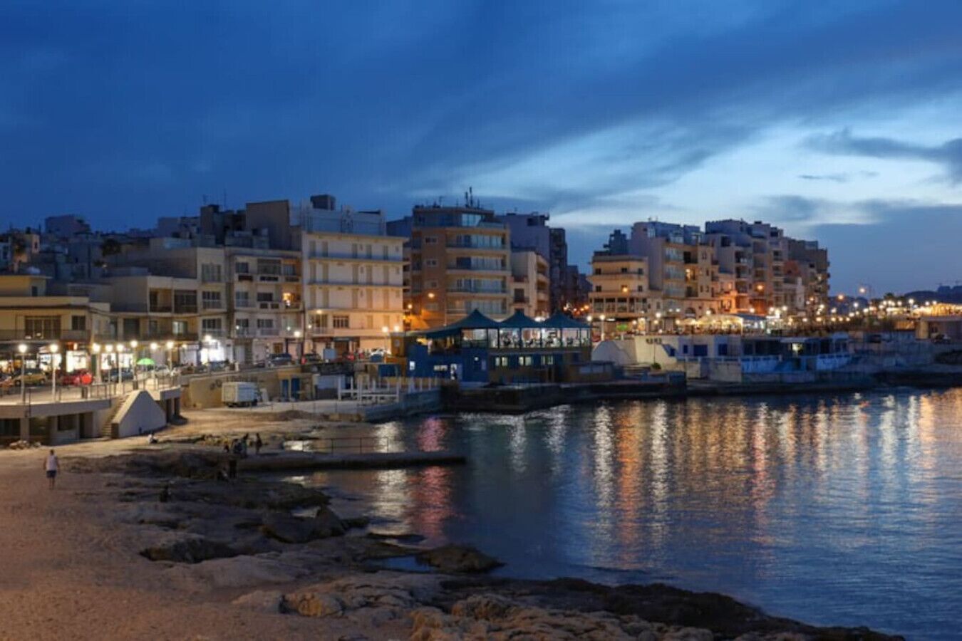 Un paysage urbain côtier au crépuscule, avec des bâtiments le long du front de mer, une plage de sable et une côte rocheuse.