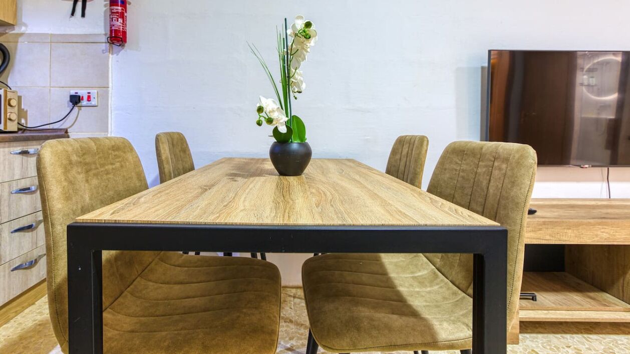Ein Esstisch mit sechs Stühlen, eine Vase mit weißen Blumen, ein Feuerlöscher und ein Fernseher an der Wand.