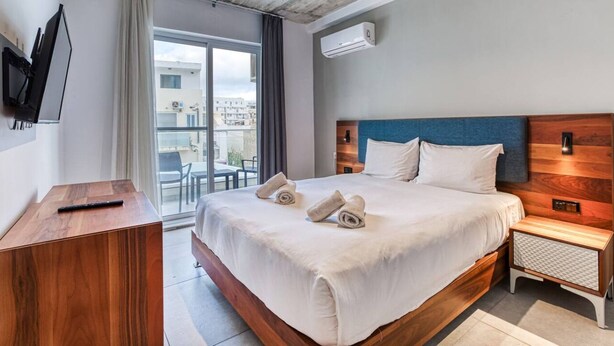 A hotel room with a bed, a TV mounted on the wall, a wooden nightstand, and a balcony with a view of buildings.