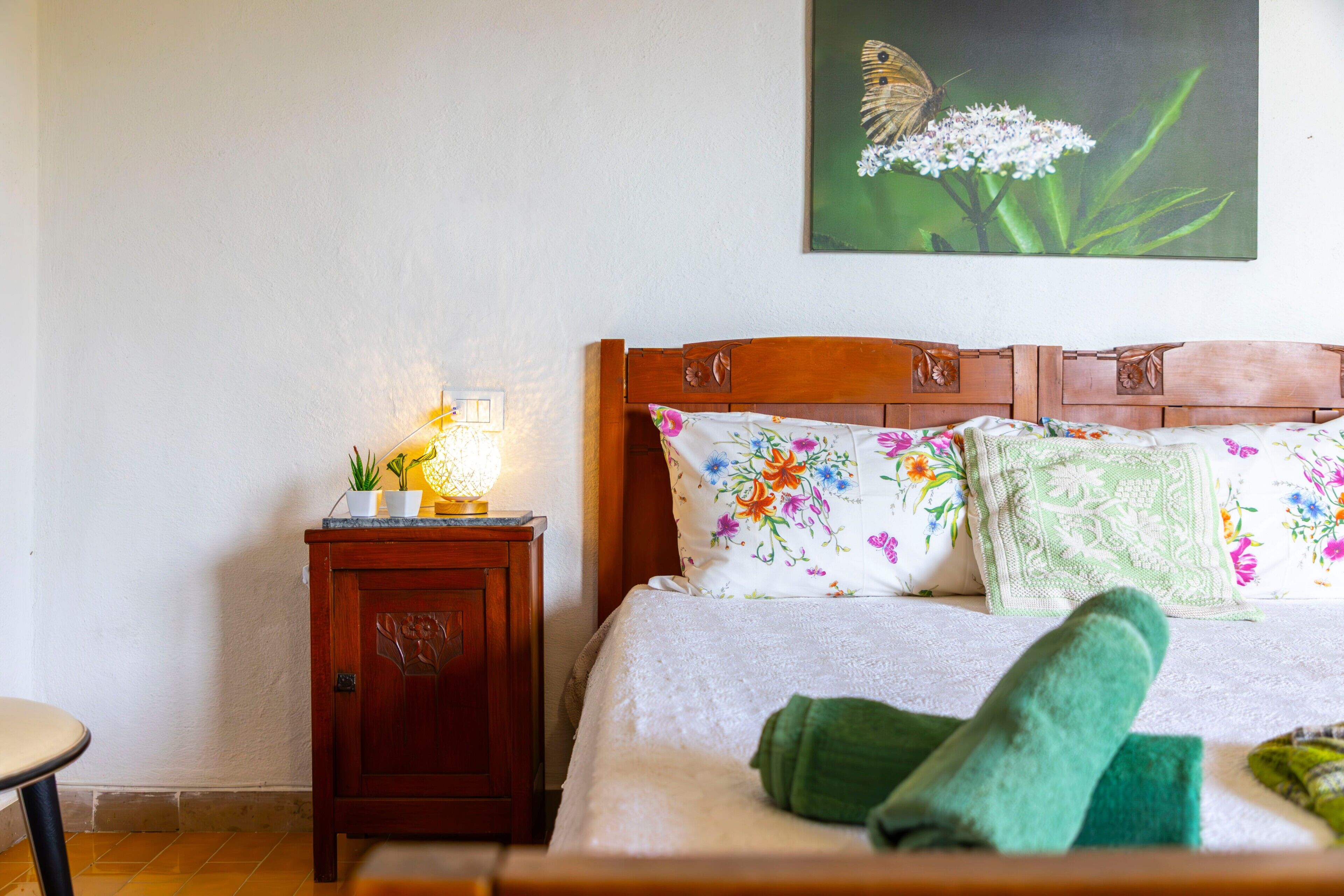 A bedroom with a wooden headboard, floral bedding, a bedside lamp, and a green towel on the bed.
