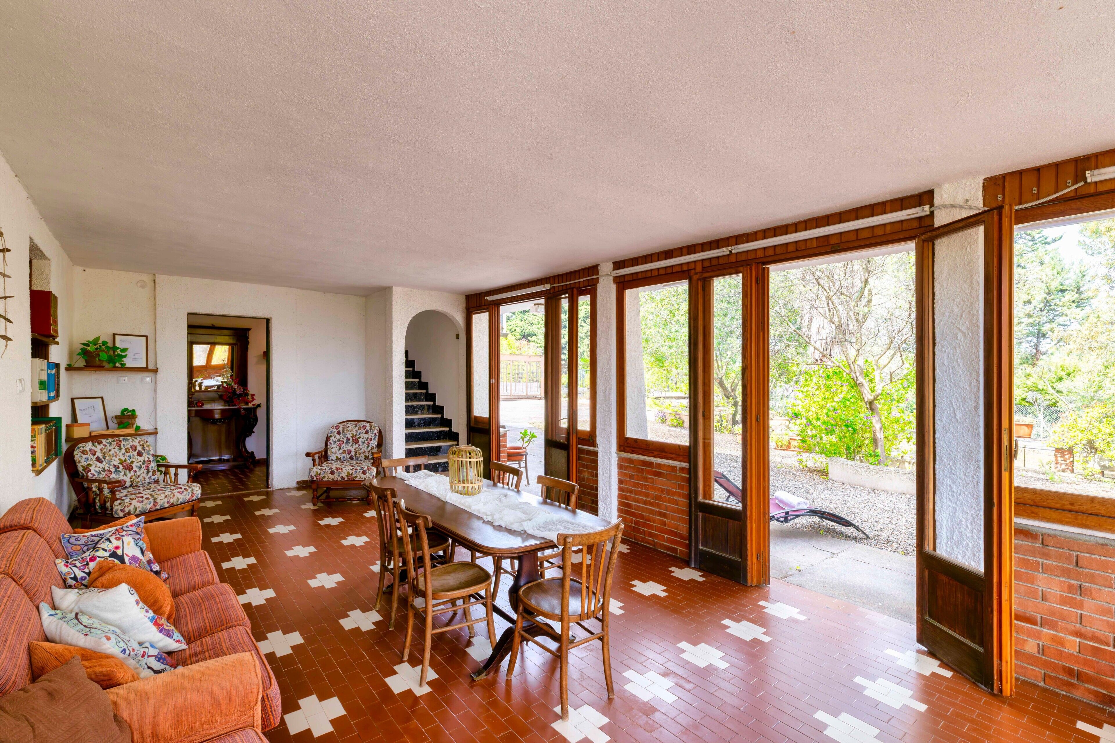 A spacious living area with a dining table, chairs, and a sofa. There is a staircase leading to an upper level, and large windows providing a view of the outdoors.