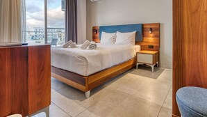Una habitación de hotel moderna con una cama grande, cabecera de madera, una mesita de noche y vistas a la ciudad.