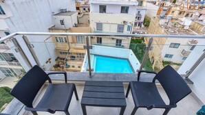Un balcón con dos sillas negras y una mesita, con vista a una piscina y una zona urbana muy poblada.