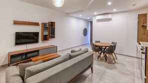 Una sala de estar moderna con un sofá gris, un televisor, estanterías empotradas, una zona de comedor con una mesa redonda y sillas, y un equipo de aire acondicionado.
