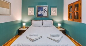 Una cama bien hecha con sábanas blancas y dos toallas dobladas al pie. Hay dos lámparas de noche sobre mesitas de madera. La habitación tiene paredes color turquesa y una puerta de madera.