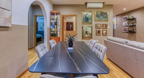 Un comedor con una mesa larga, sillas con cojines, un sofá y una estantería con cuadros enmarcados.