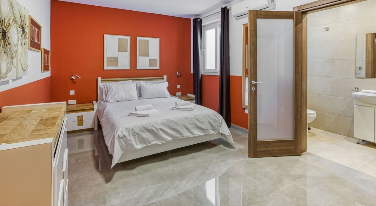 A hotel room with a bed, a nightstand, a bedside table, a bathroom with a toilet and sink, and a door leading to another room.