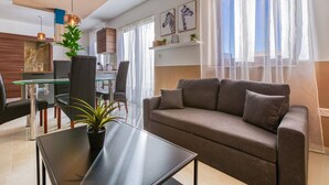 A modern living room with a sofa, a glass dining table, and a coffee table. There is a kitchen area in the background with a stove and cabinets.