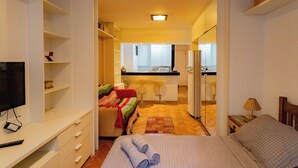 A hotel room with a bed, a TV, a desk, a chair, a sofa, and a kitchen area visible through an open door. - SUH Flamengo 01 Praia Studio Dreams (Rio de Janeiro)
