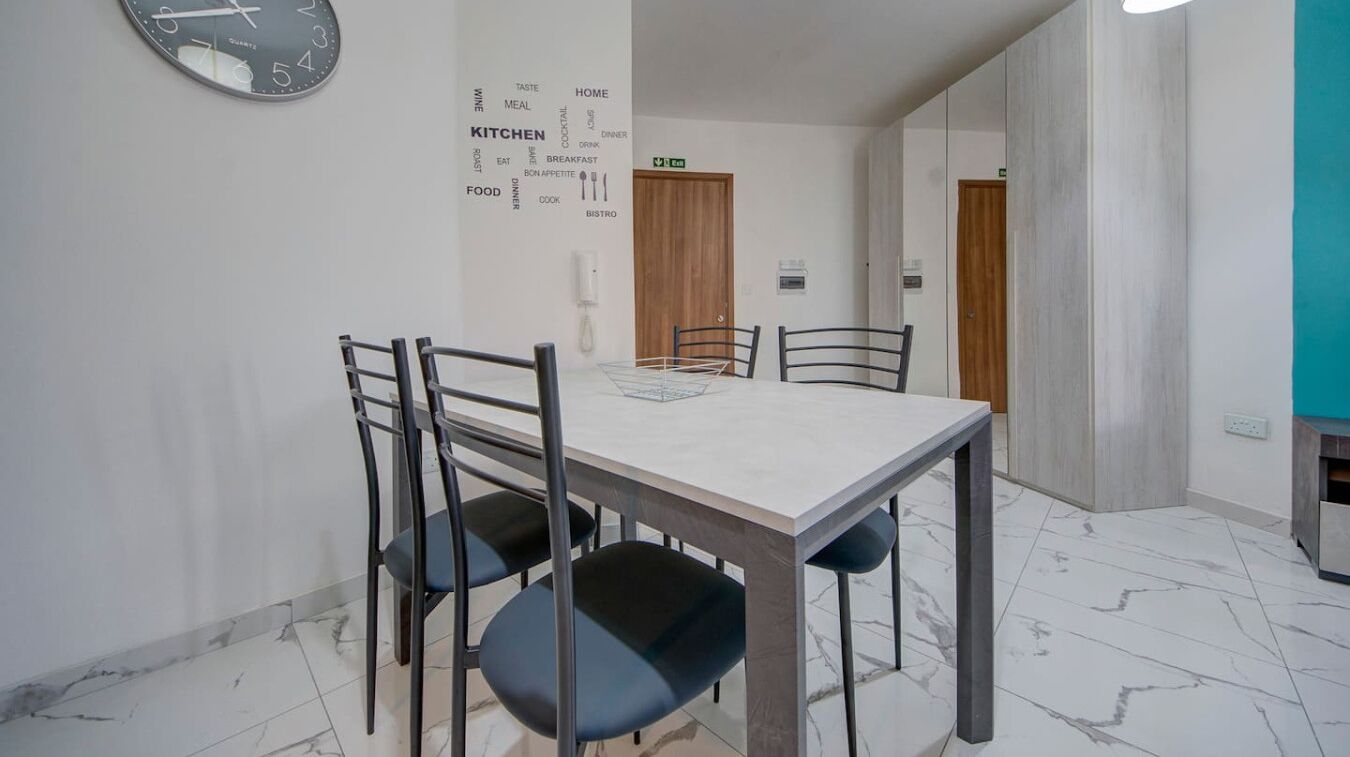 Un comedor con piso de mármol, una mesa blanca y cuatro sillas. Se observa un reloj de pared y una nube de palabras relacionada con comida y cenas.