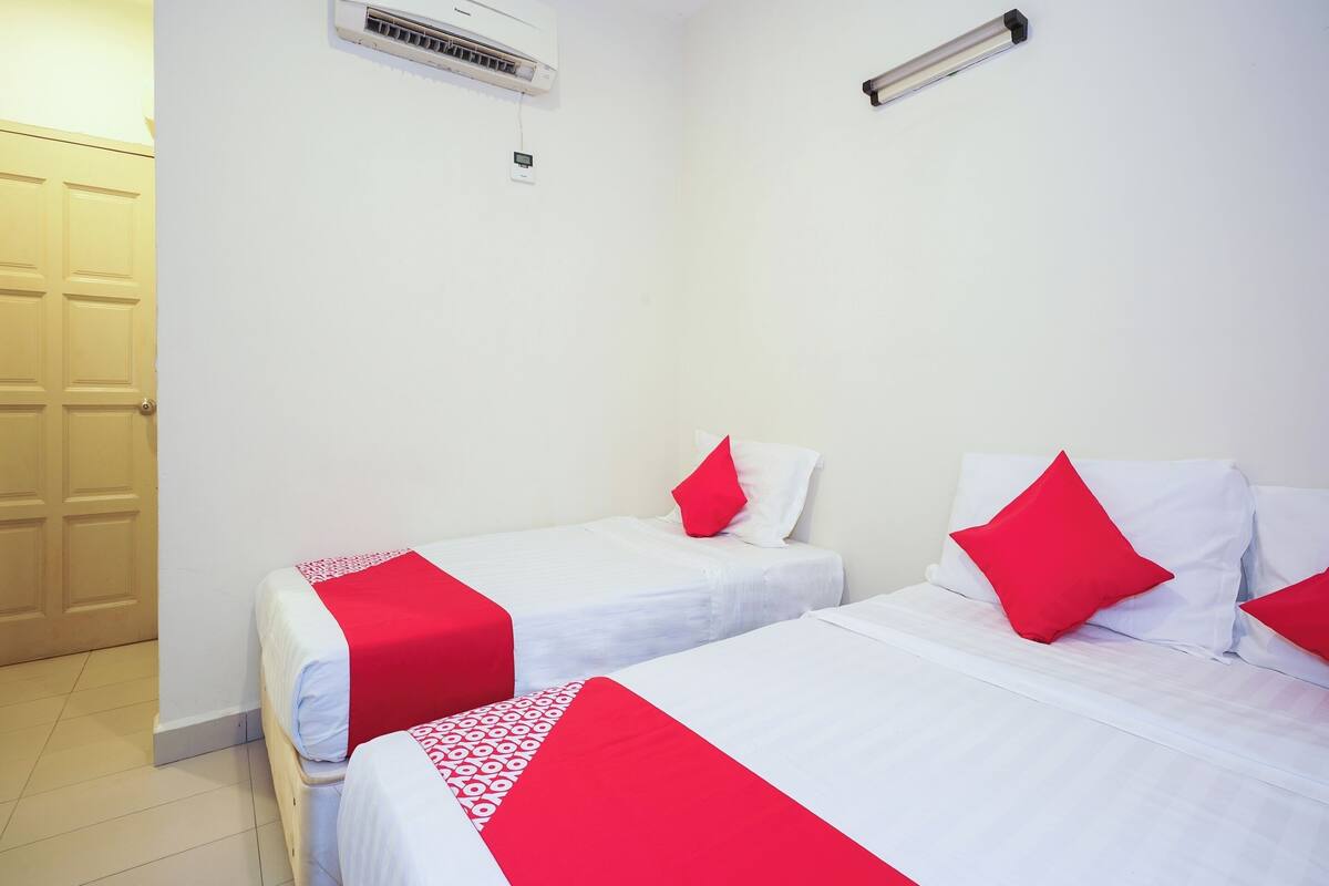 Une chambre d’hôtel avec deux lits, chacun agrémenté de draps blancs et de touches de couleur rouge, un climatiseur blanc et une porte en bois.