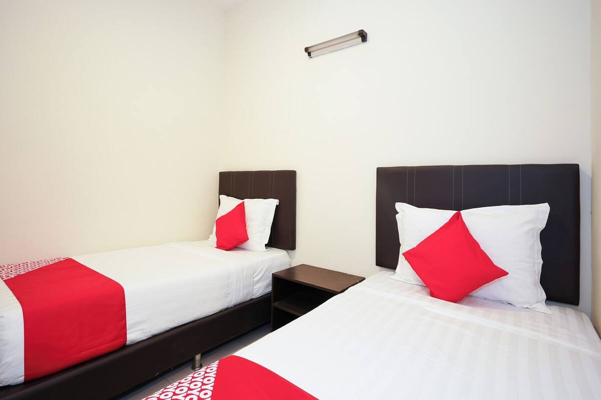 Une chambre d’hôtel avec deux lits simples, chacun recouvert d’une courtepointe rouge et blanche, dotée d’une tête de lit sombre et d’une petite table de chevet.