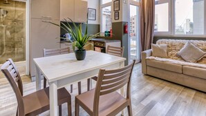 A modern living room with a sofa, a dining table with chairs, a potted plant, and a kitchen area with cabinets and a window.