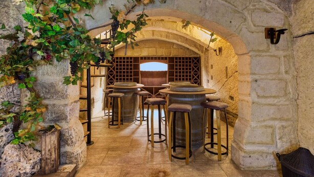 A rustic stone interior with a vaulted ceiling, a central table with stools, and a wooden barrel.
