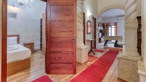 A hotel room with a wooden door, a bed, a nightstand, a chandelier, and a view of the gym area.
