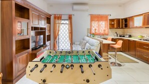 A room with a foosball table, a sofa, and a kitchen area with wooden cabinets and a window with curtains.