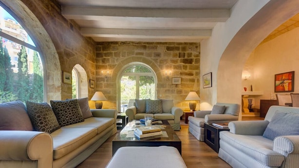 A spacious living room with a stone wall, a large arched window, and a cozy seating arrangement including a sofa, armchairs, and a coffee table. - Stunning Villa with private pool & large terrace! (Balzan)