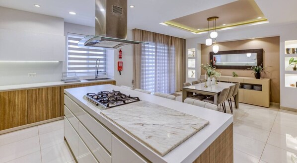 Cozinha moderna com ilha central, sala de jantar com mesa e cadeiras e sala de estar com sofá e televisão.