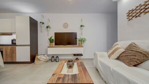 A modern living room with a TV mounted on a white wall, a wooden coffee table, a sofa with cushions, and a kitchen area in the background.