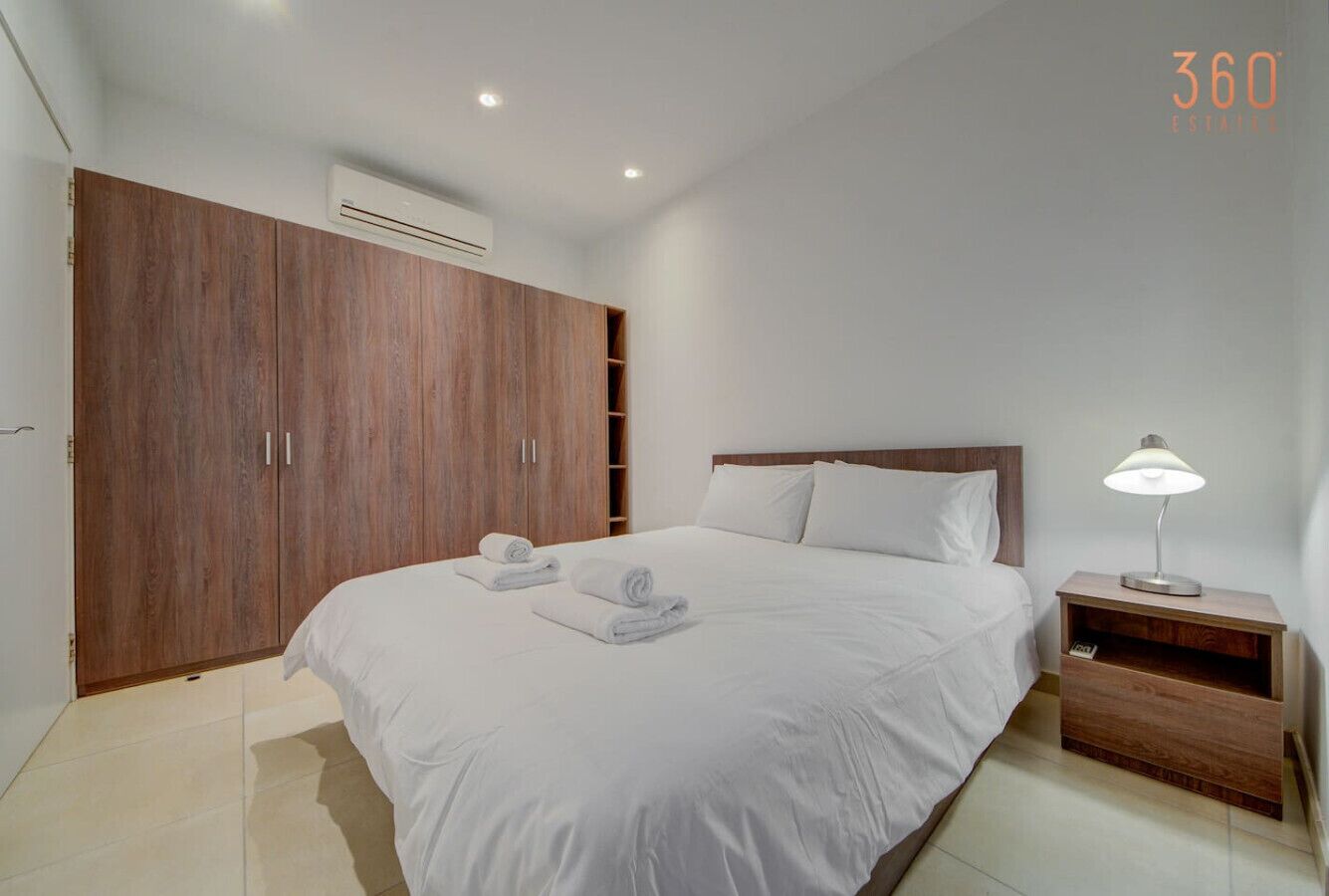 Een slaapkamer met een grote kledingkast, een bed met witte lakens en een nachtkastje met een lamp.