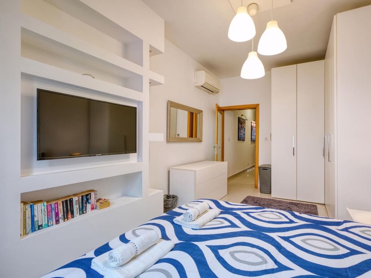 A modern hotel room with a large bed, a flat-screen TV mounted on the wall, a bookshelf with books, a wardrobe, and a mirror.