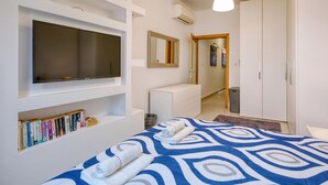 A modern hotel room with a large bed, a flat-screen TV mounted on the wall, a bookshelf with books, a wardrobe, and a mirror. - Stylish St. Julian’s 1BR home, Cozy & Central! (St. Julian`s)