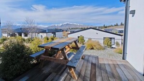Eine Holzterrasse mit Picknicktisch, umgeben von Grünflächen und Blick auf moderne Gebäude und Berge.