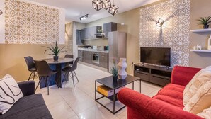 Un salón moderno con un sofá rojo, una zona de comedor con mesa y sillas, una cocina al fondo y un televisor sobre un mueble.