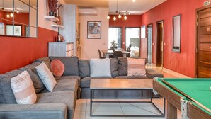Una sala de estar moderna con mesa de billar, sofá gris con cojines estampados y un comedor al fondo.
