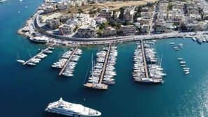 An aerial view of a marina with numerous boats docked, surrounded by a coastal cityscape with buildings and a clear sky.