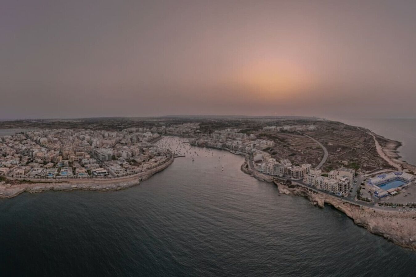 An aerial view of a coastal city with a prominent curved harbor, dense urban development, and a large resort area with multiple buildings.